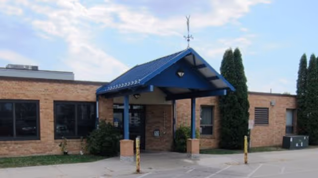 Brick facility entrance with a blue covered canopy and surrounding shrubs.