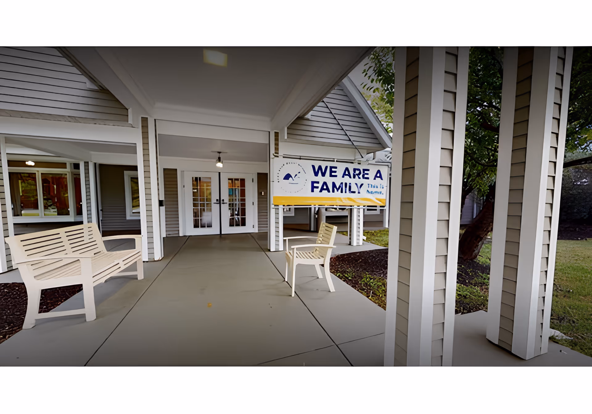Covered entrance porch of a senior living facility with benches, double doors, and a sign reading "WE ARE A FAMILY".