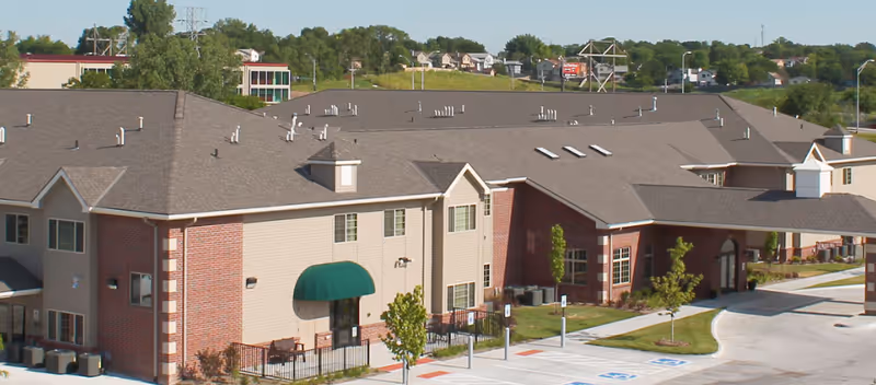 Two-story senior living building with brick and beige siding, a green awning over an entrance, a covered drive-up entry, and adjacent parking.