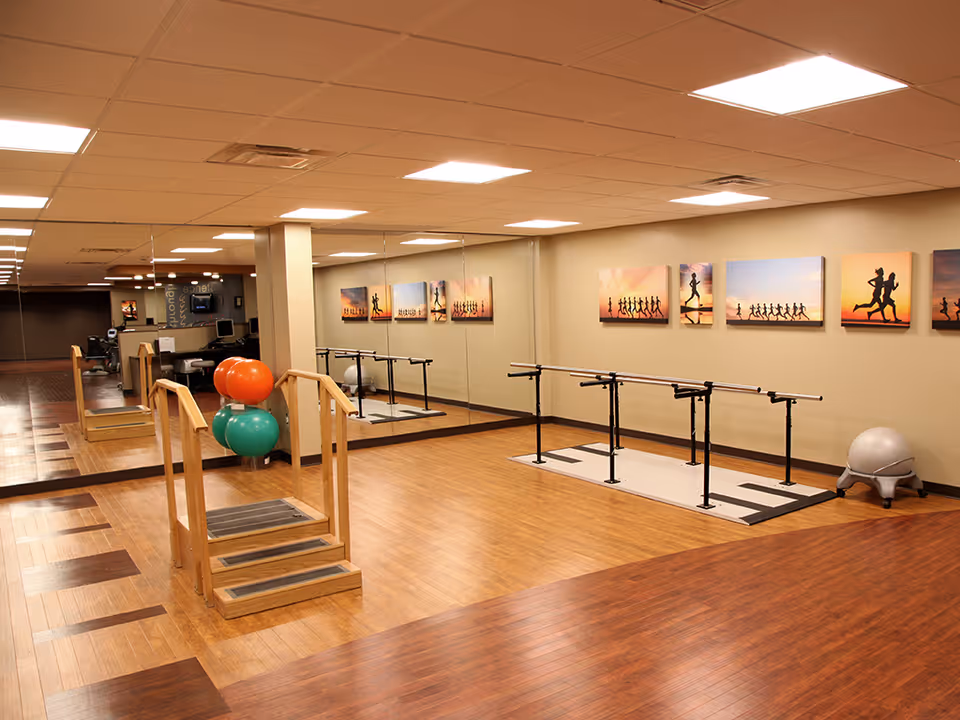 A spacious physical therapy or exercise room with wooden flooring, parallel bars for walking exercises, a small set of stairs with handrails, exercise balls, and a large wall mirror reflecting the room. The walls are decorated with framed pictures of people running at sunset.