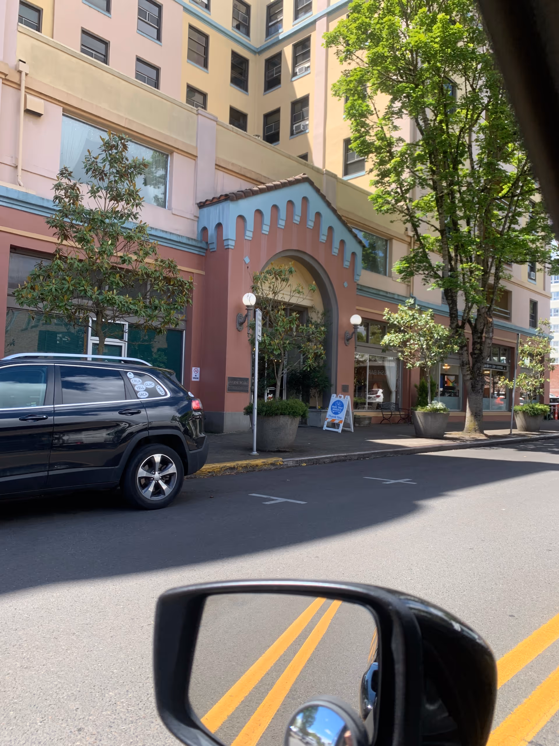Exterior view of a pastel-colored building with arched entrance, trees, parked car, and sidewalk.