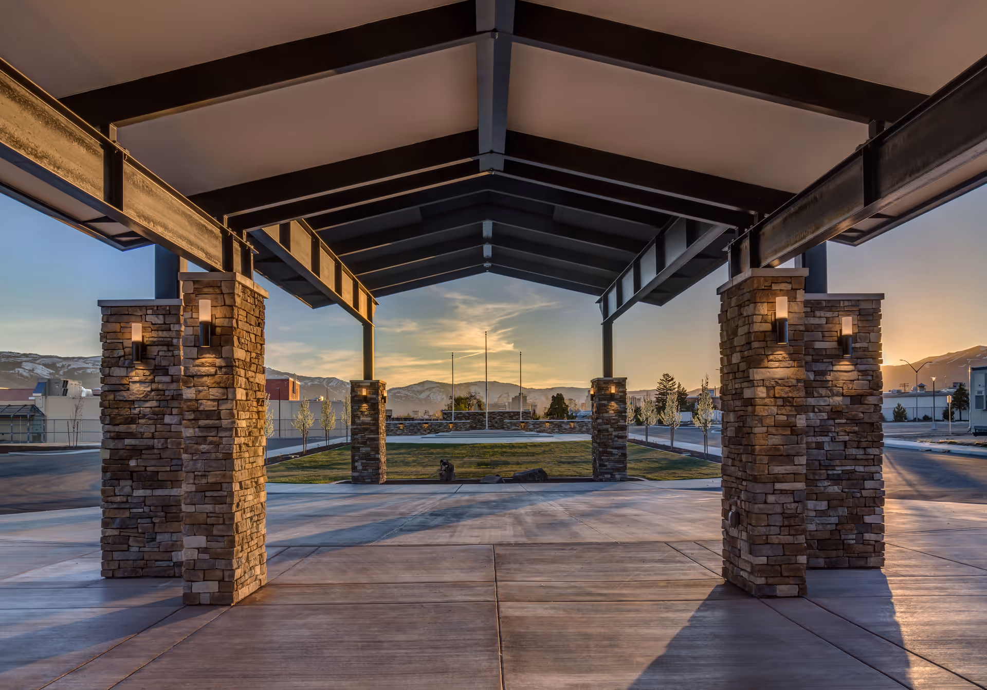 Covered entrance canopy with stone pillars and exposed steel beams overlooking a landscaped drive and distant mountains at sunset.