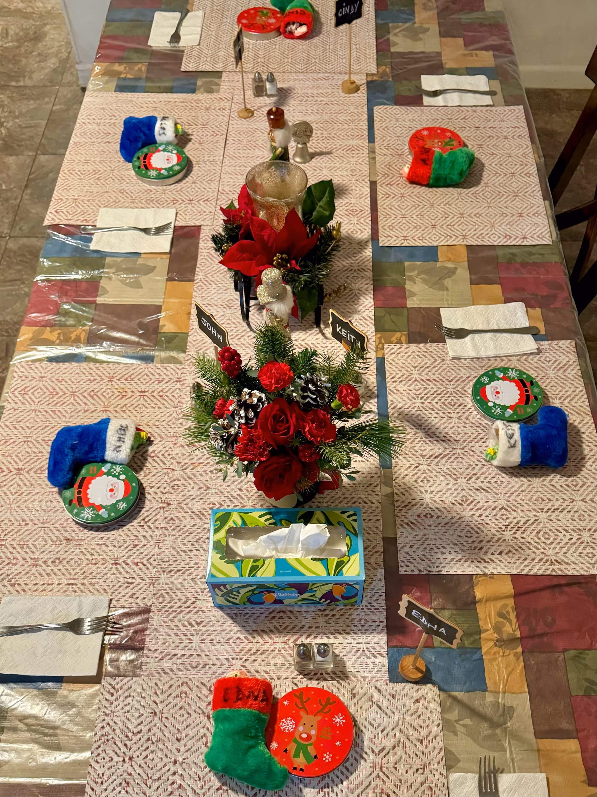 A dining table set for six with festive Christmas decorations including small stockings, plates with Santa and reindeer designs, and floral centerpieces with red flowers and pine cones. Each place setting has a napkin and fork, and there are name tags indicating seating arrangements.