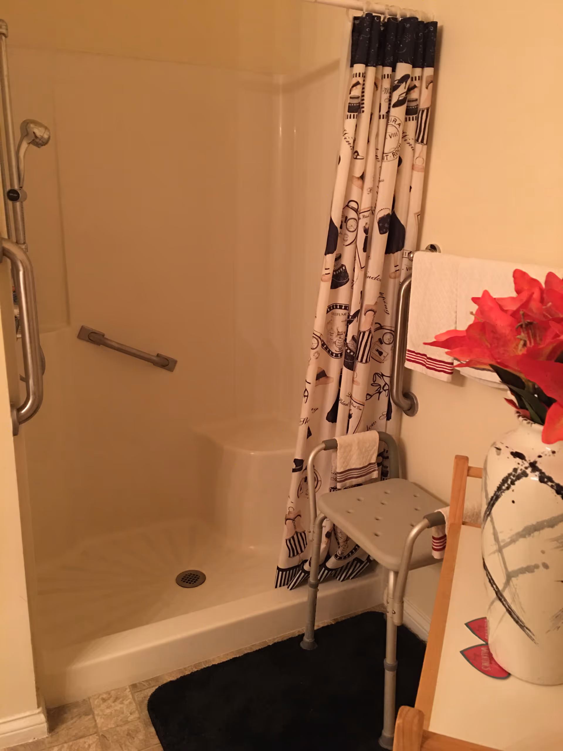 A bathroom shower area with a white shower stall, a patterned shower curtain, and grab bars installed on the walls. There is a gray shower chair placed inside the shower area. A towel hangs on a grab bar outside the shower. A vase with red flowers is visible on a nearby surface.