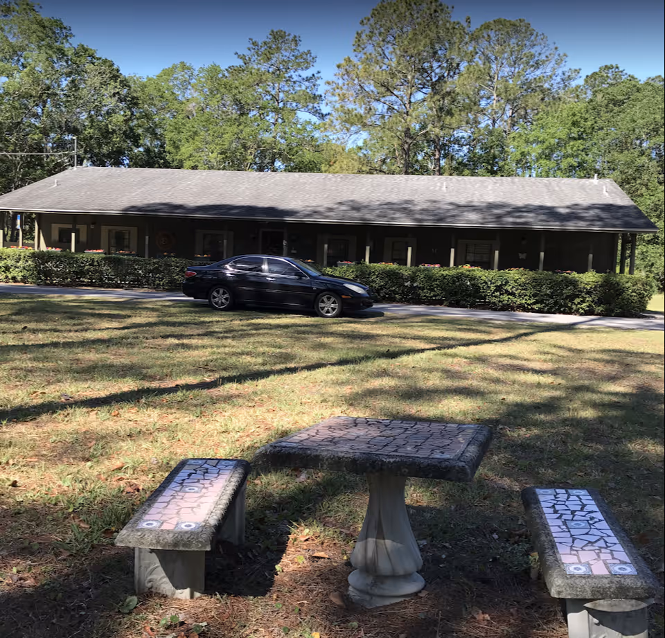 Outdoor scene showing a single-story building with a gray roof and a porch partially obscured by bushes. A black car is parked on a driveway in front of the building. In the foreground, there is a stone table with mosaic tile patterns and two matching benches on a grassy area with trees in the background under a clear blue sky.