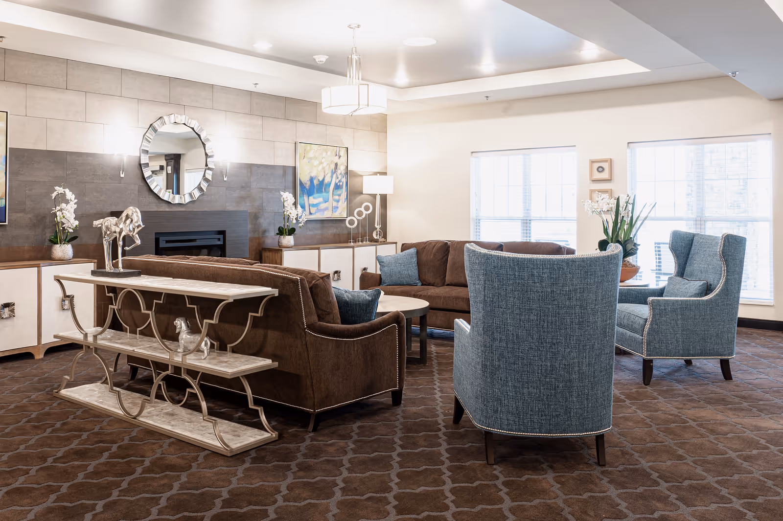 A well-lit living room with brown sofas and blue armchairs arranged around a circular coffee table. The room features a decorative fireplace with a round mirror above it, two large windows letting in natural light, and various decorative items including plants and artwork on the walls.