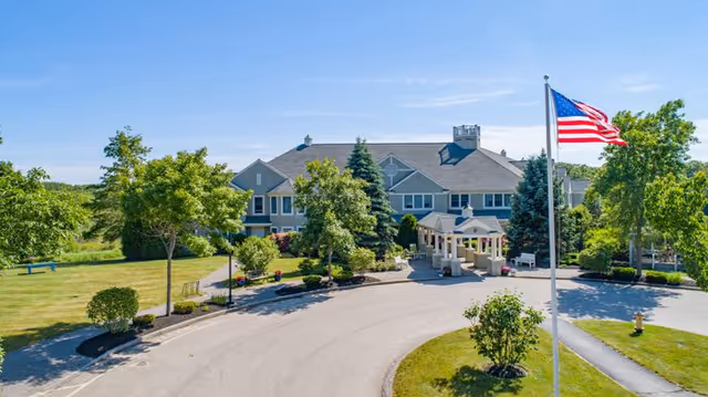 Front exterior view of The Landing at Saco Bay facility with a circular driveway, landscaped greenery, trees, and an American flag on a flagpole in the foreground under a clear blue sky.