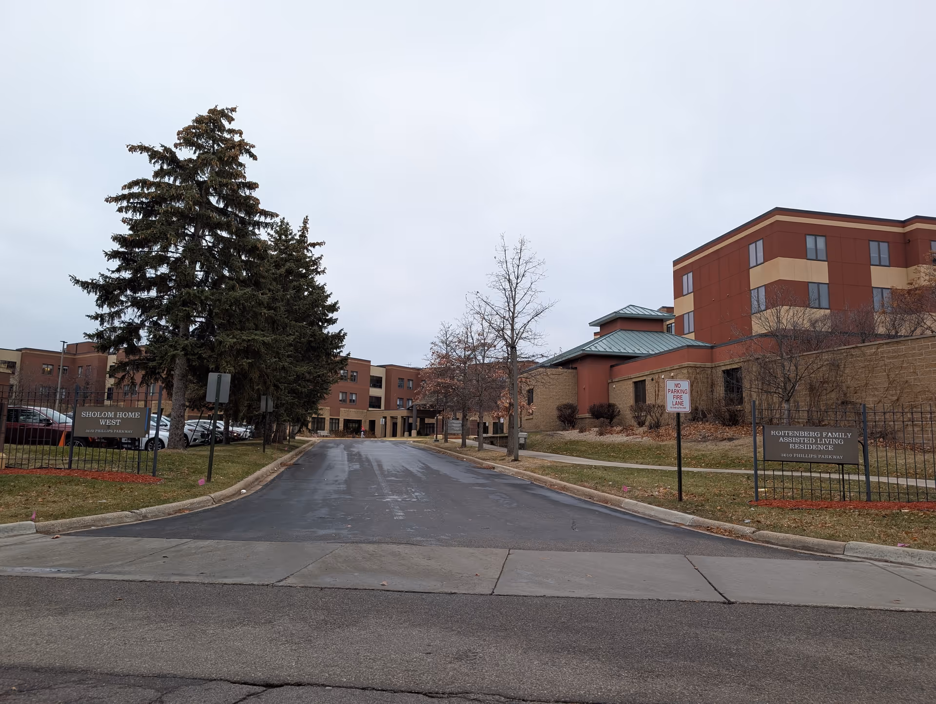 Front driveway and entrance of a multi-story brick senior living facility with trees, signs, and a fenced lawn area.