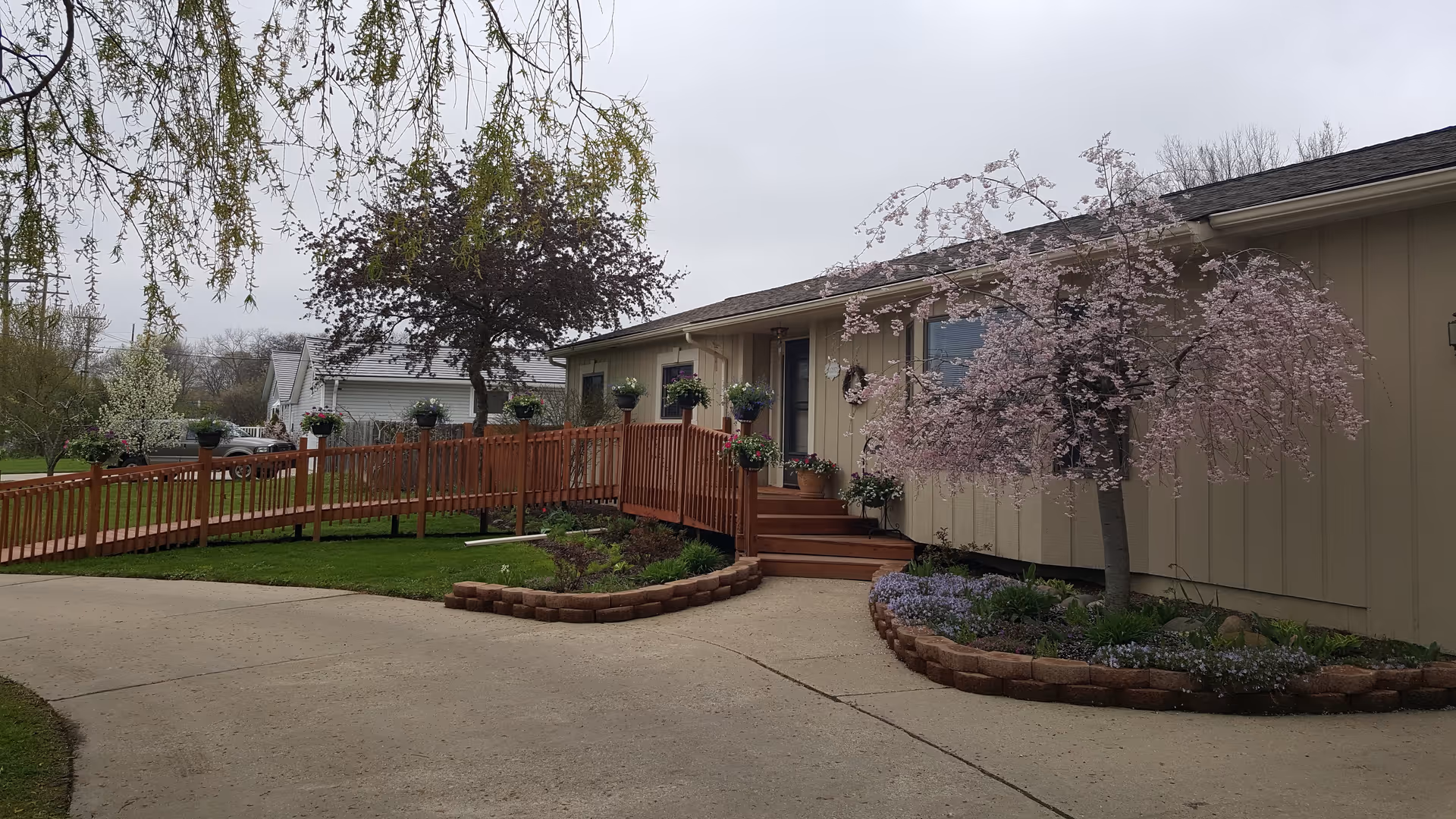 Exterior view of a single-story building with beige siding, a wooden ramp and stairs leading to the entrance, surrounded by landscaped flower beds and trees with pink and purple blossoms under an overcast sky.