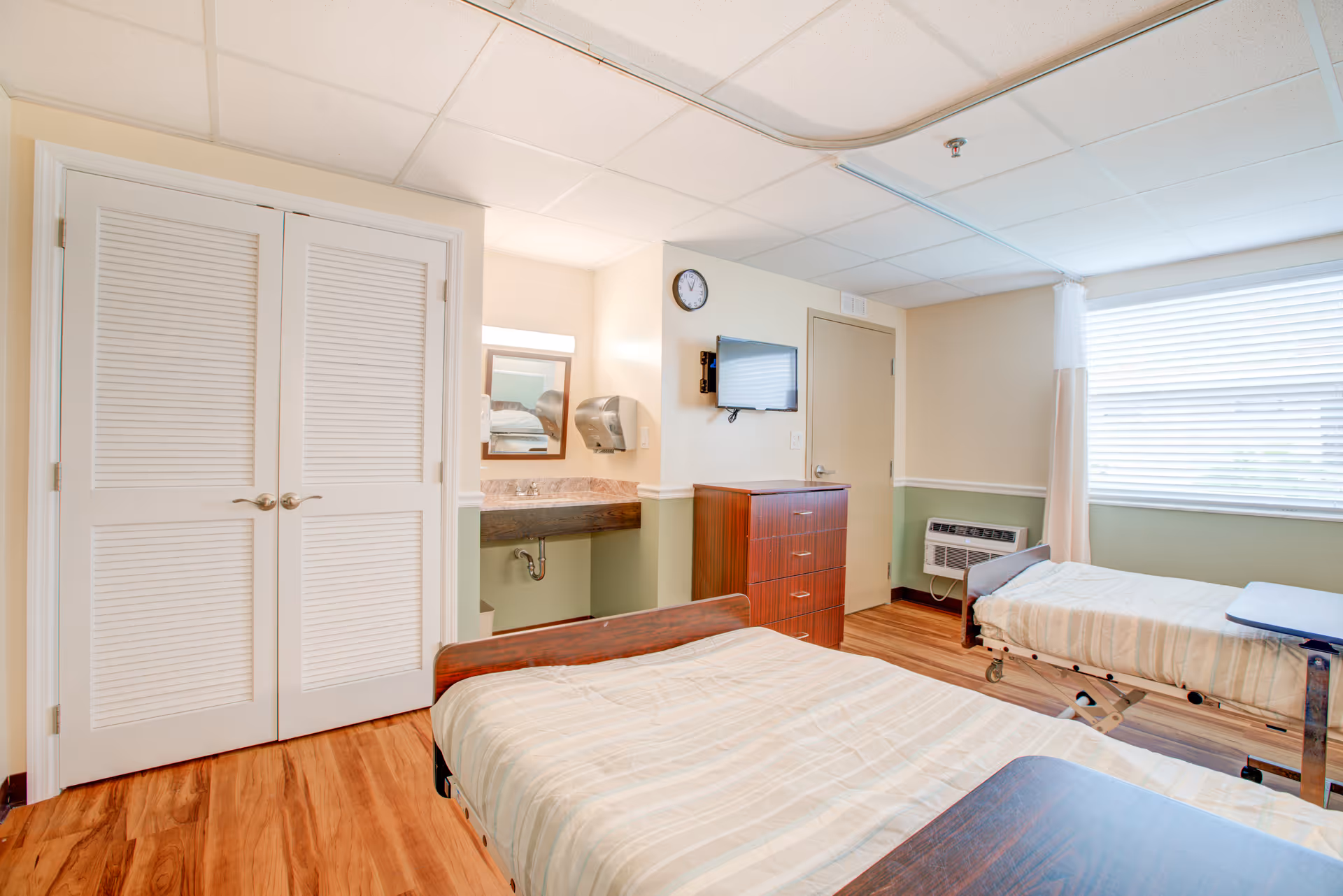 Two beds in a shared patient room with a dresser, sink area, and window.