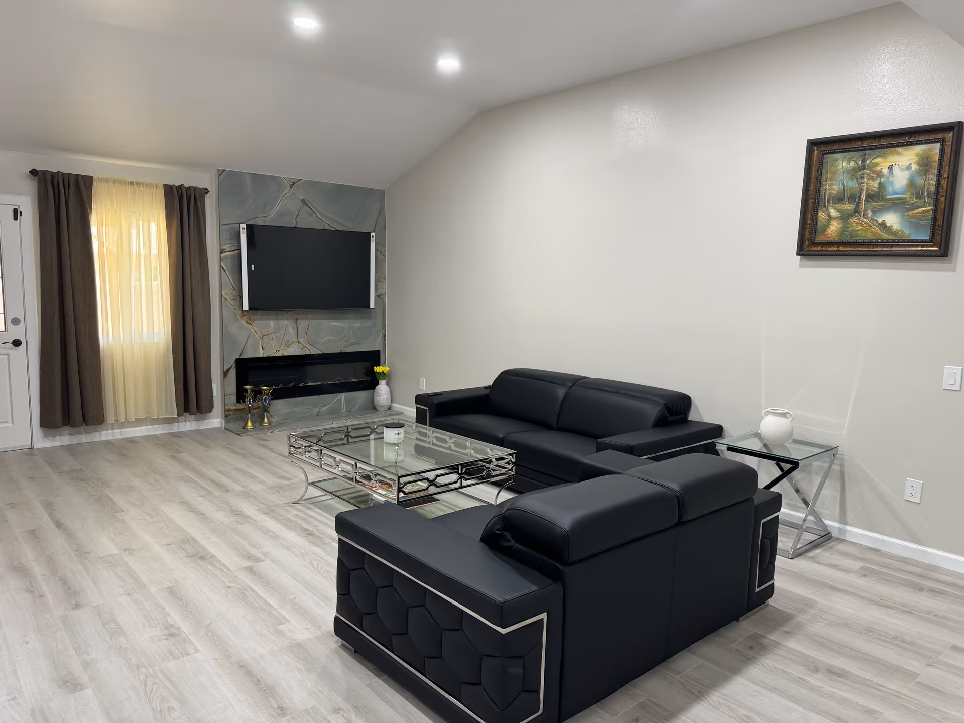 A spacious modern living room with black leather sofas, a glass coffee table, a wall-mounted TV above a tiled fireplace, light wood floors and a framed painting.