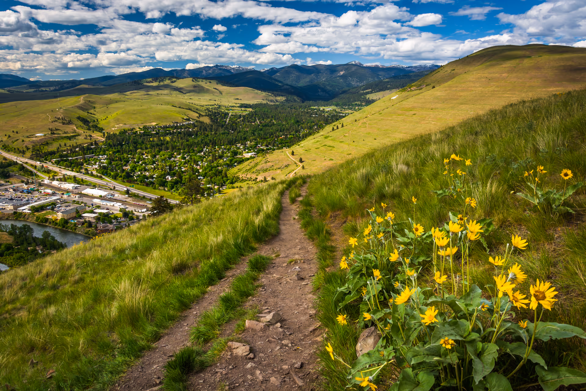 A dirt hiking trail winds through grassy hills toward a valley town beneath mountains.