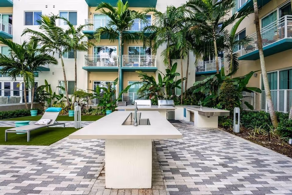 Outdoor courtyard with a long white kitchen island, lounge chairs, palm trees and surrounding apartment balconies.