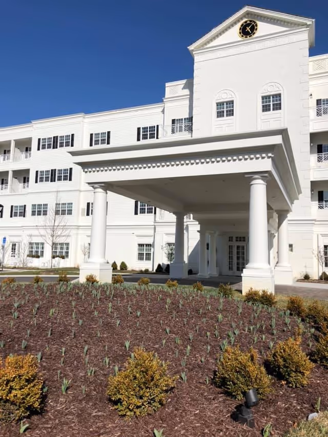 Front entrance of a white multi-story senior living building with a porte-cochère supported by columns, a clock tower, and landscaped planting beds in front.