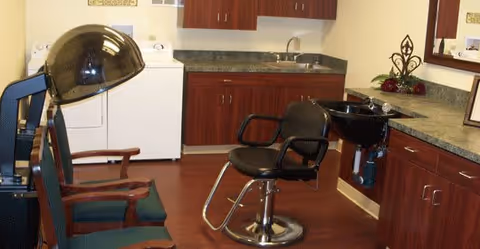 Interior view of a salon area within a facility, featuring a black salon chair in front of a black hair washing sink, two green cushioned chairs, wooden cabinets with a countertop, a mirror, and a washing machine in the background.