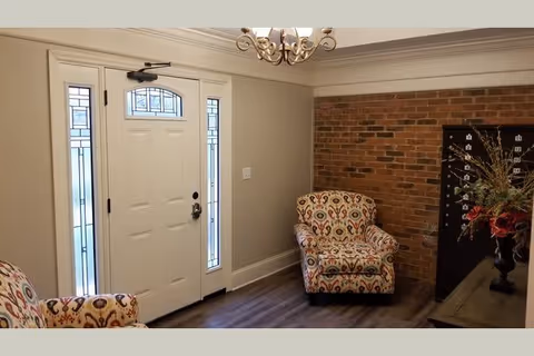 Interior entryway with a white front door flanked by narrow windows, two patterned armchairs, a brick accent wall, and a chandelier.