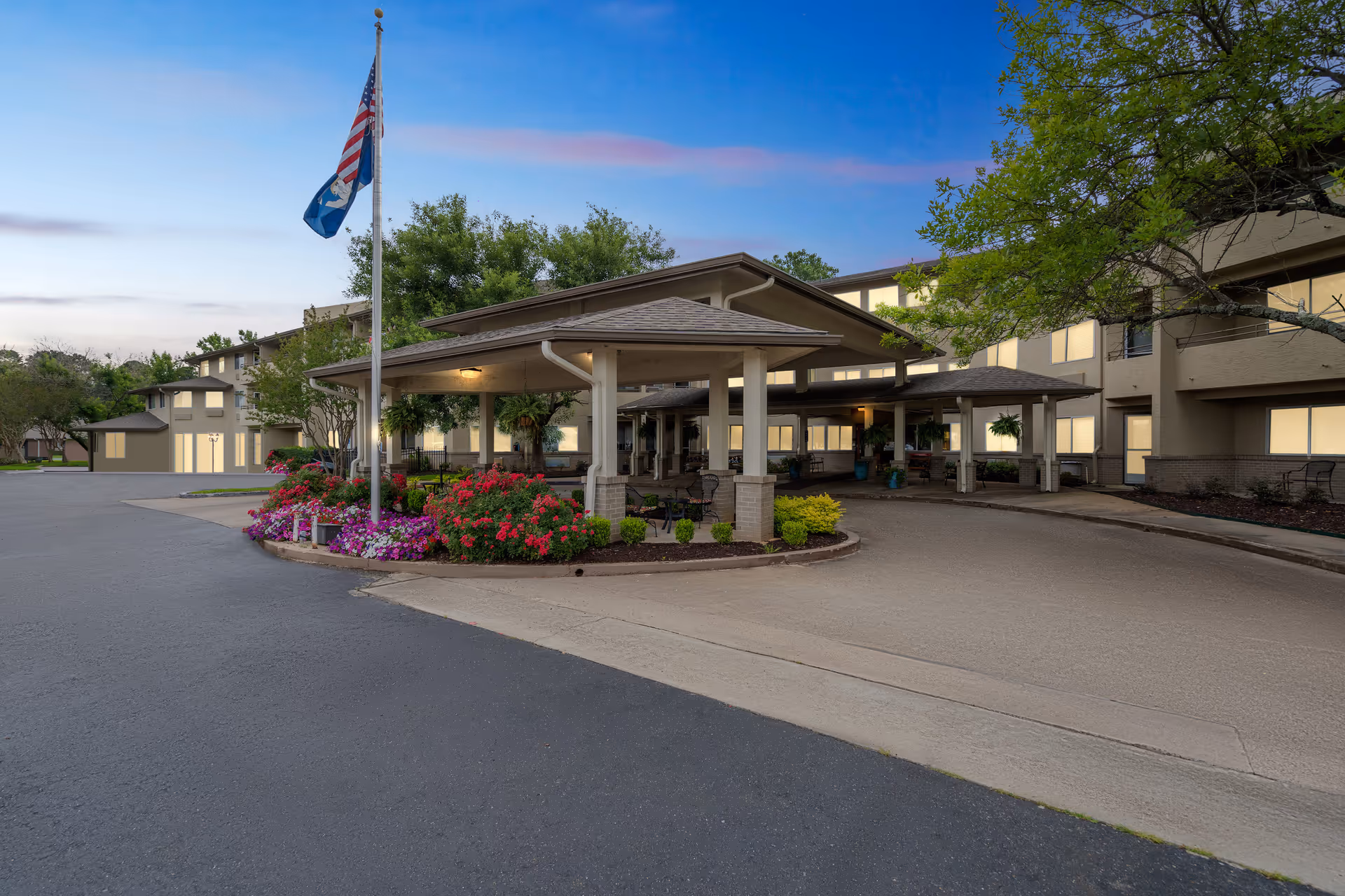 Exterior view of Holiday Summerfield Estates senior living facility at dusk, showing the main entrance with a covered drop-off area, landscaped flower beds, an American flag and a state flag on a flagpole, and a multi-story building in the background.