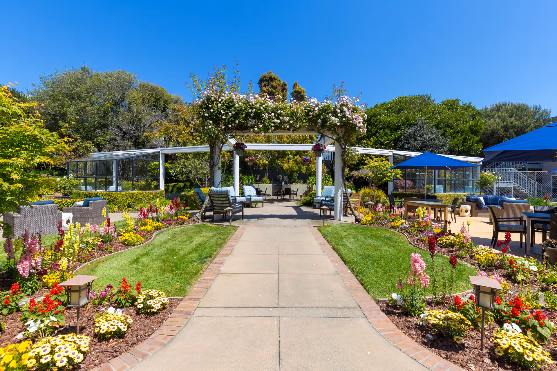 Pathway leading to a flower-covered pergola and outdoor seating area surrounded by manicured gardens and umbrellas under a blue sky.