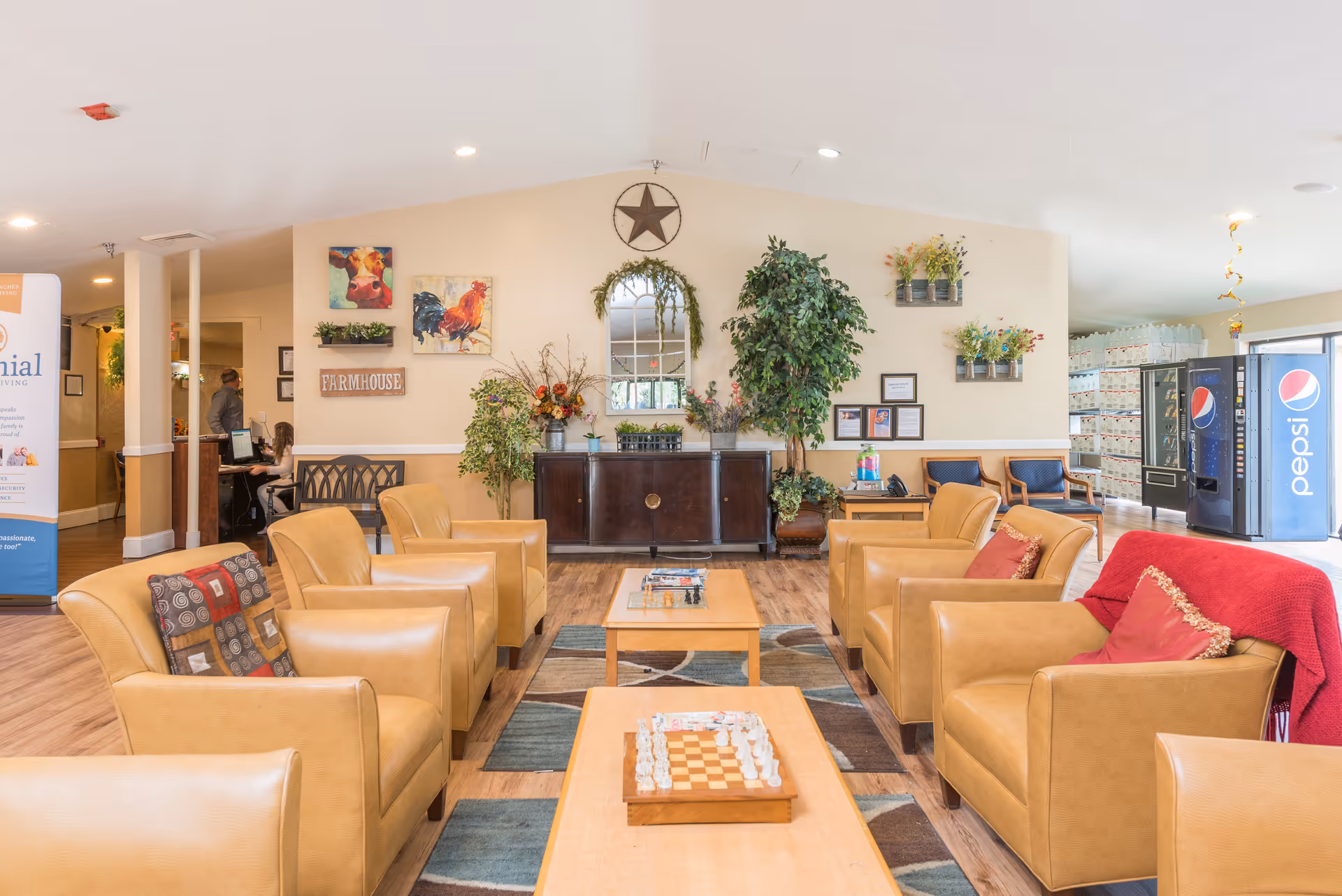 Bright senior living facility lobby with tan leather chairs, coffee tables, plants, and vending machines.
