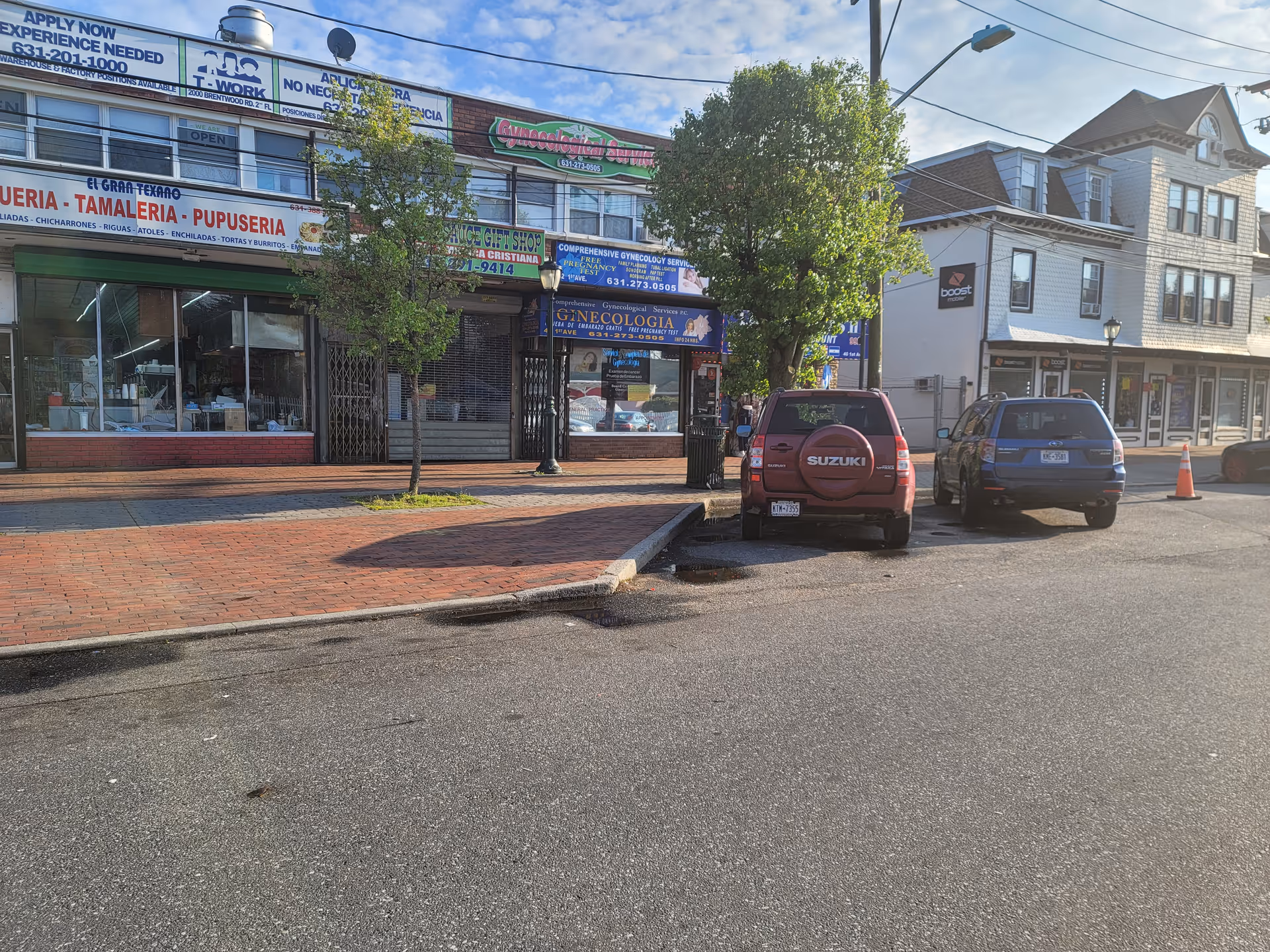 Urban storefronts line a street with parked cars and trees.