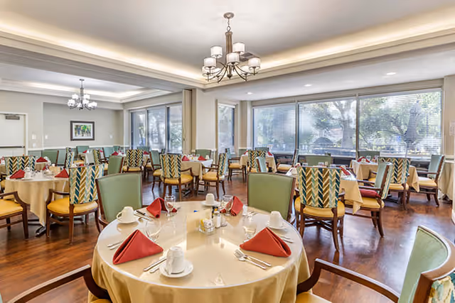 A bright dining room with multiple round tables covered with beige tablecloths, each set with white cups, plates, silverware, and red folded napkins. The room has large windows letting in natural light, wooden floors, and green and patterned chairs. Ceiling lights and a chandelier provide additional lighting.
