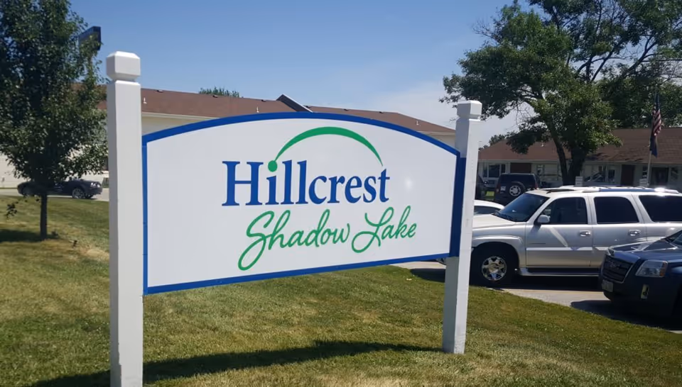 White sign on two posts reading "Hillcrest Shadow Lake" on a grassy lawn with cars and a building behind it.