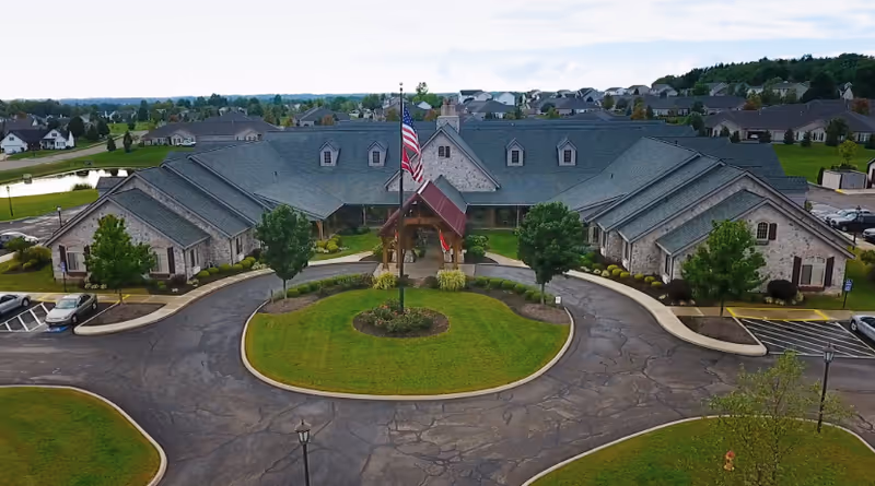 Aerial view of The Inn At University Village, showing a large, single-story building with a gray roof and stone exterior. The building has a covered entrance with an American flag and another flag in front. There is a circular driveway and landscaped greenery surrounding the entrance. Residential houses and a pond are visible in the background.