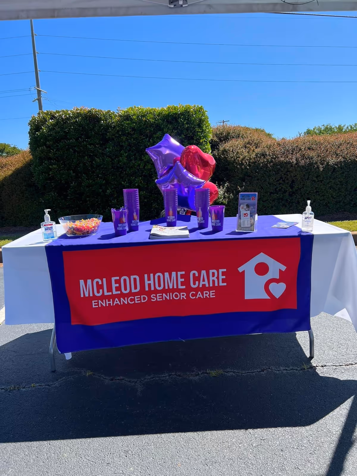 Outdoor promotional table with McLeod Home Care banner, balloons, and hand sanitizer.