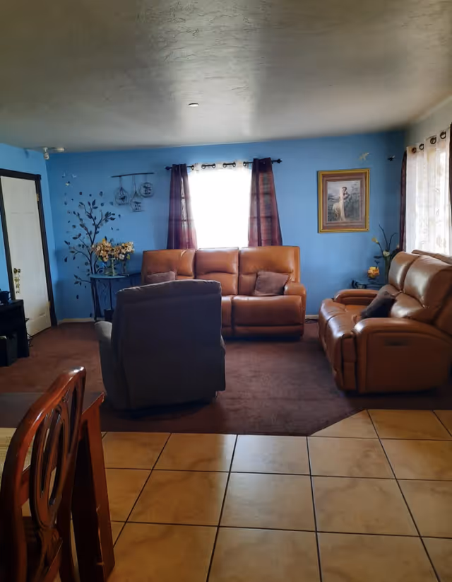 Cozy living room with tan leather sofas and recliners, a gray armchair, blue painted walls, curtains over a central window, and tiled floor transitioning to carpet.