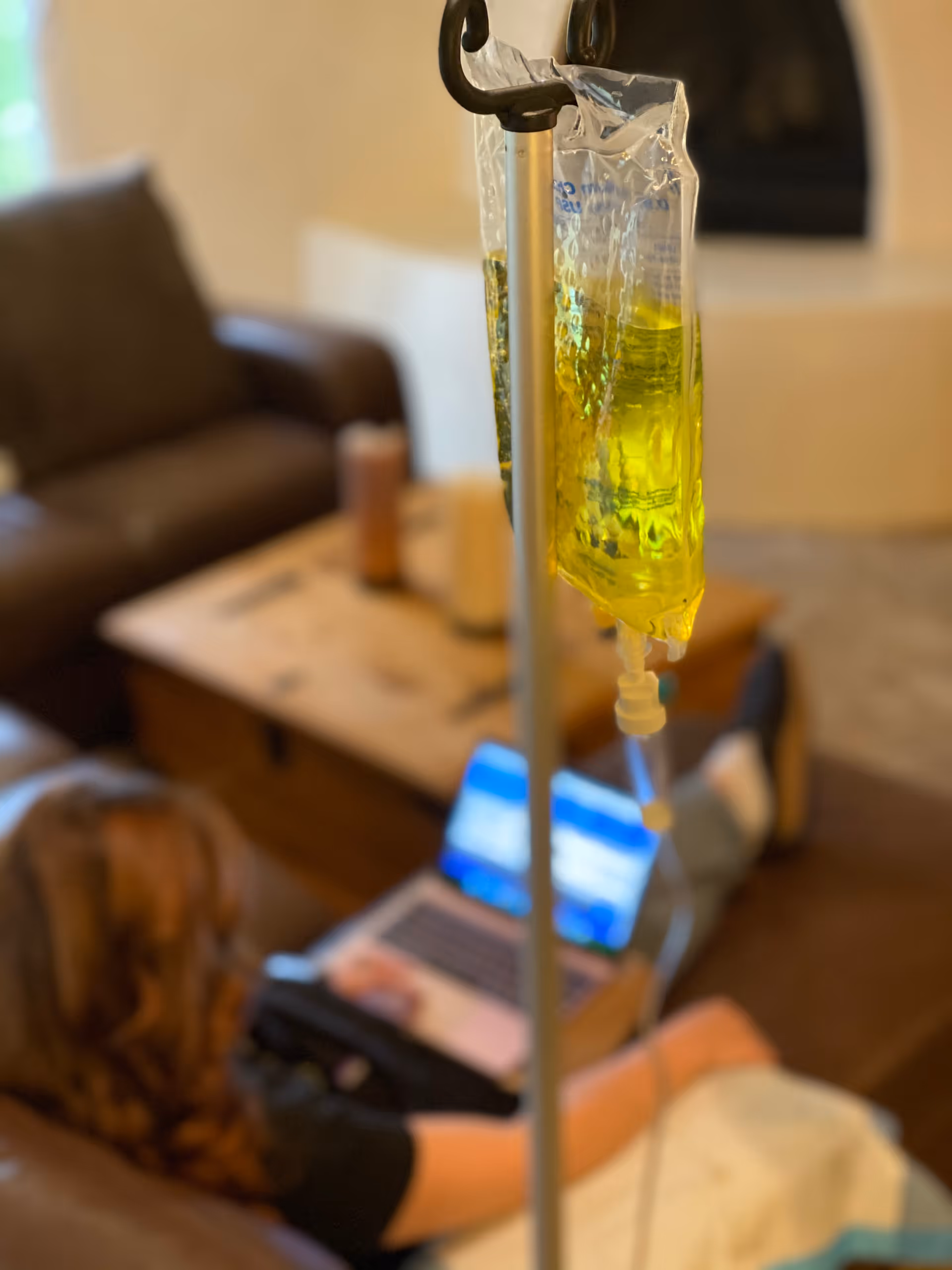 IV drip bag hanging in foreground with a resident using a laptop in a living room.