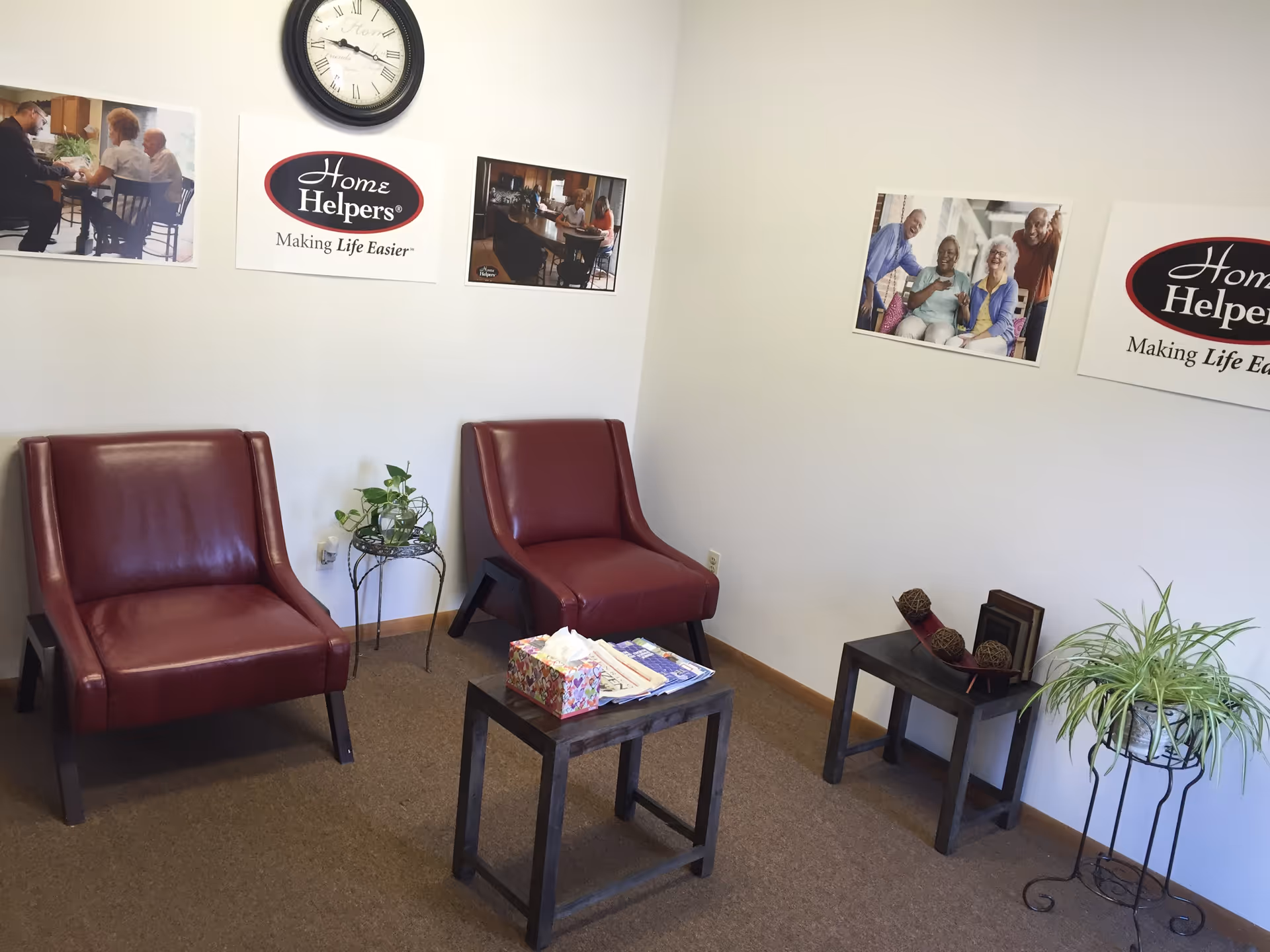 A small waiting lounge with two burgundy chairs, side tables, plants, and wall posters.