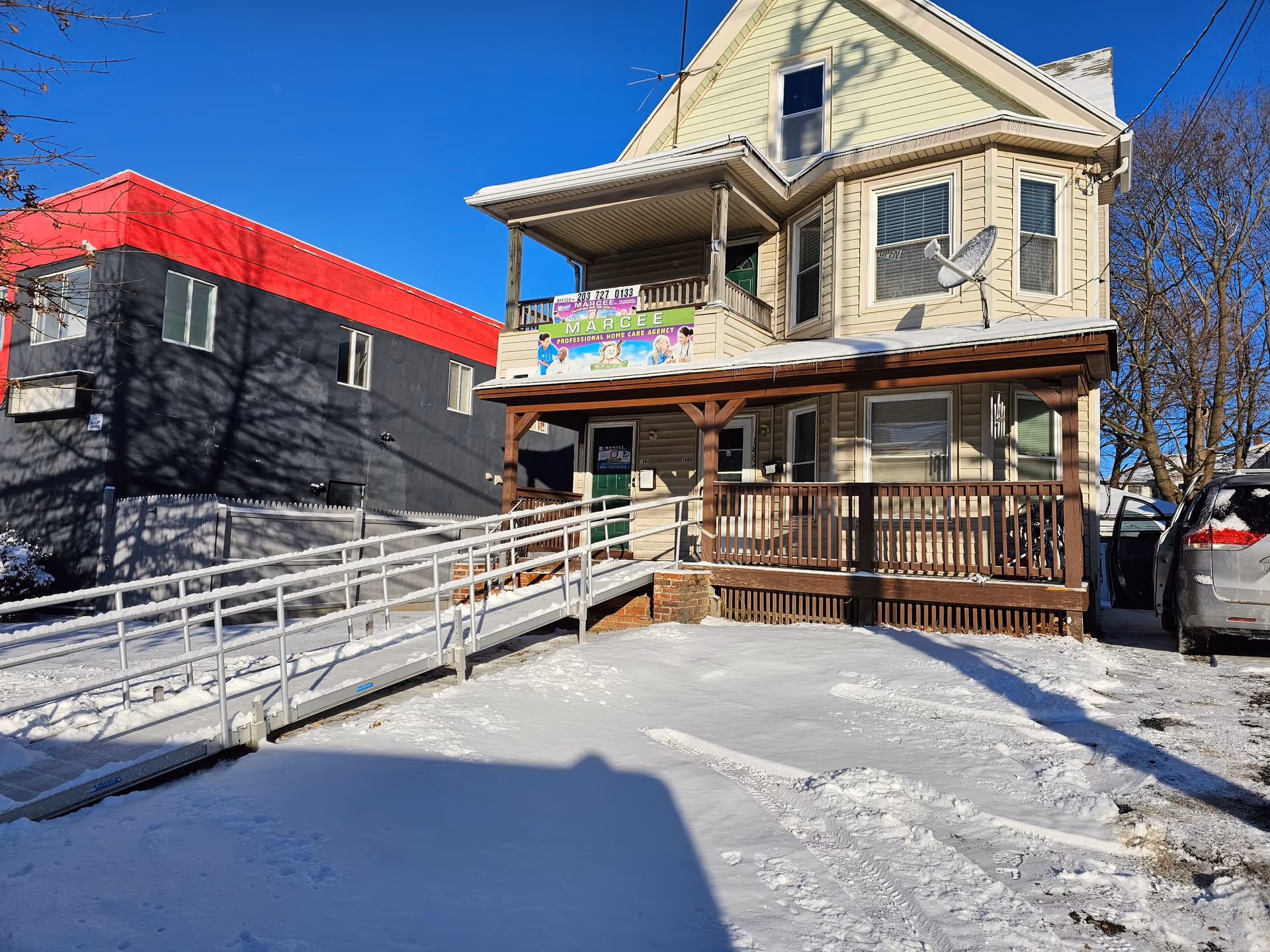 Two-story beige house with front porch, banner, and wheelchair ramp in snowy lot.