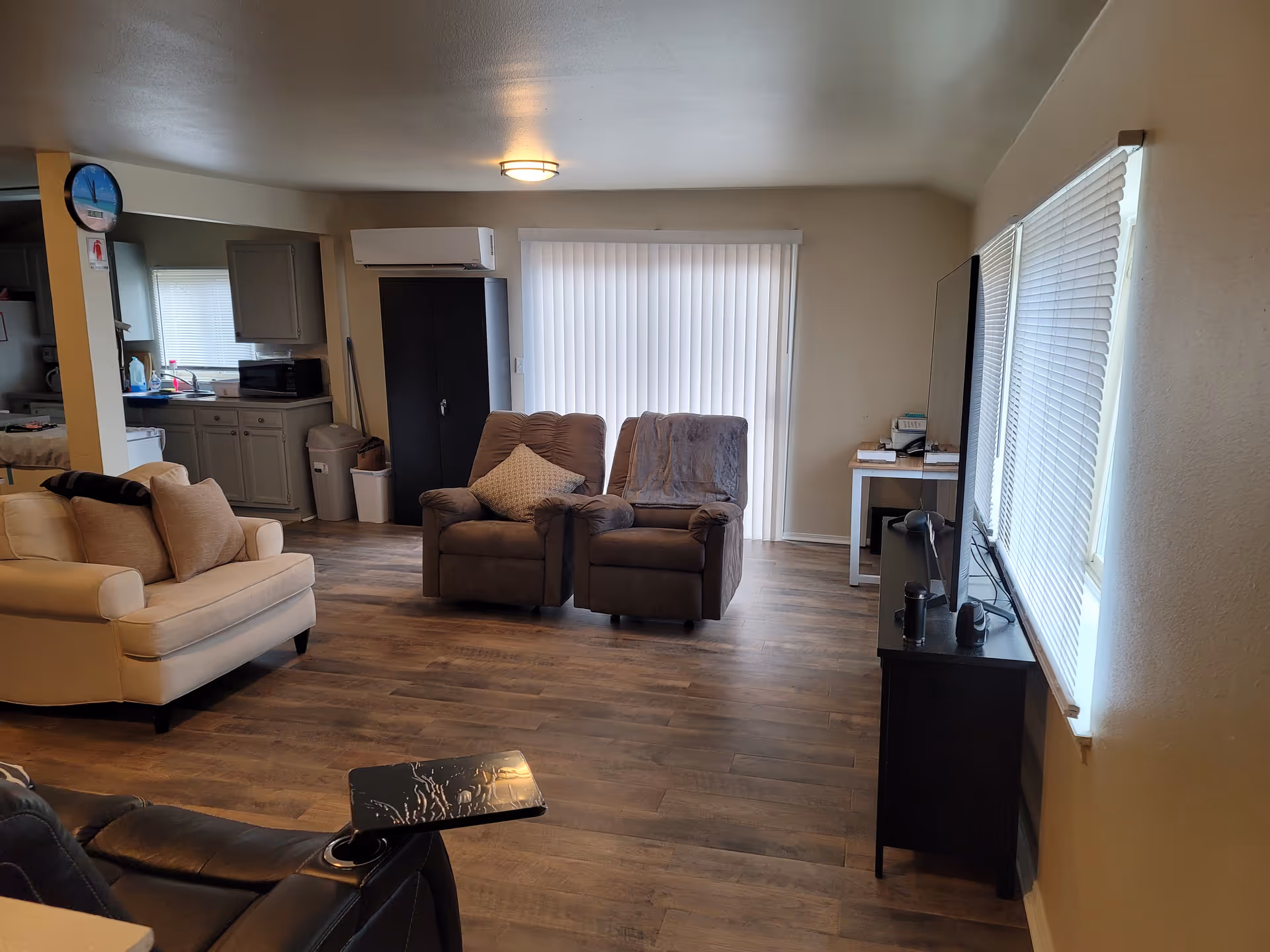 Living room with beige sofa, two recliners, TV, and kitchenette visible in background.