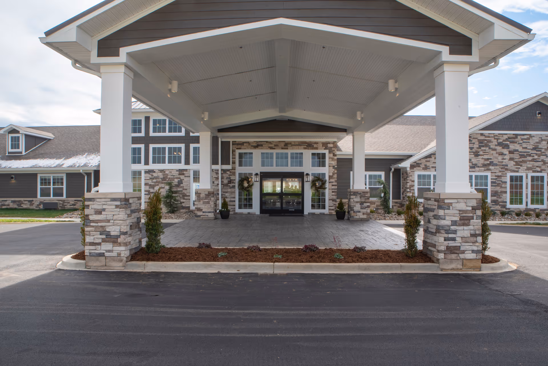 Covered entrance of a senior living facility with stone pillars and a paved driveway. The building exterior features stone and siding with multiple windows and double glass doors decorated with wreaths.