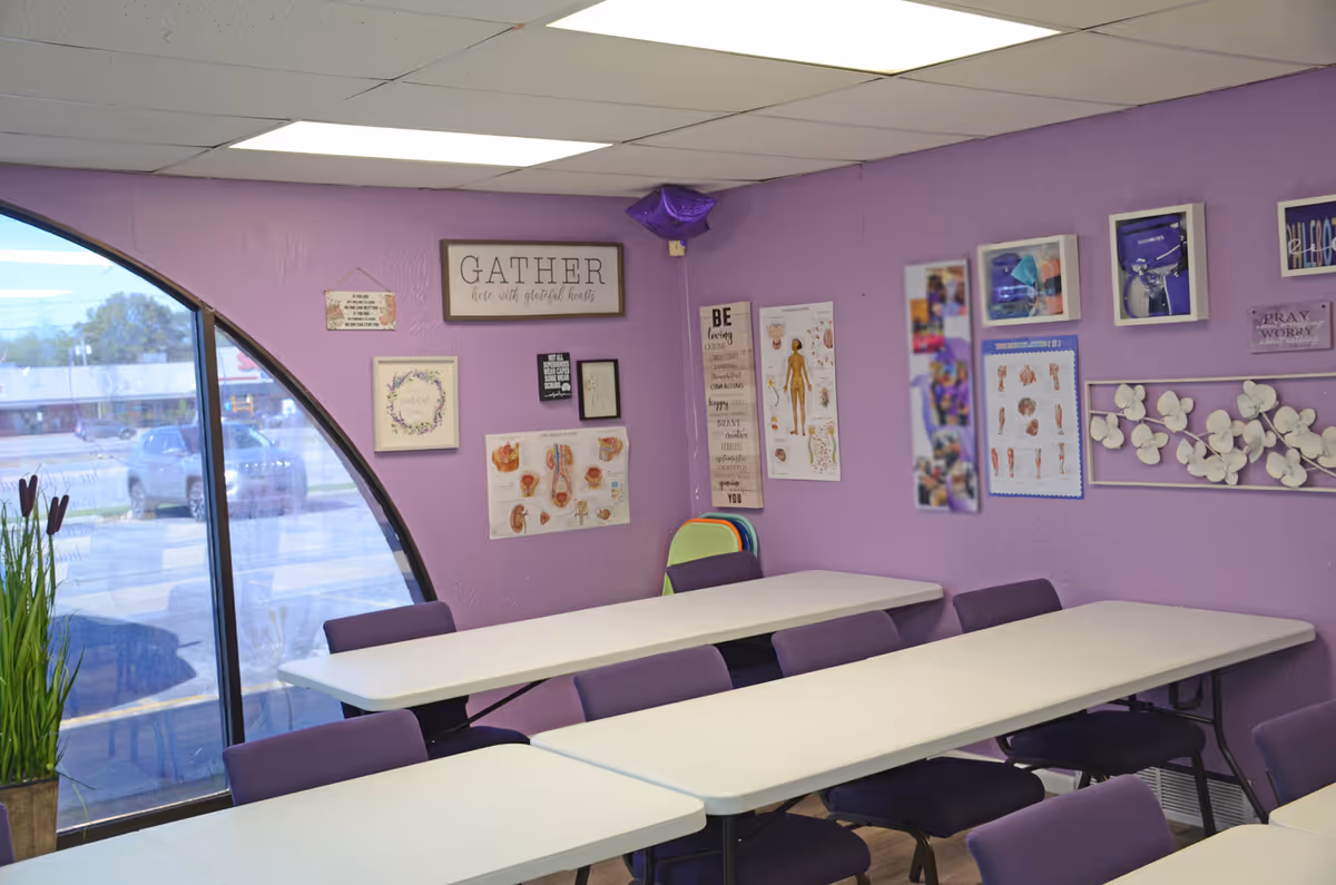 Purple-walled classroom-style room with long tables, chairs, posters, and a curved window.
