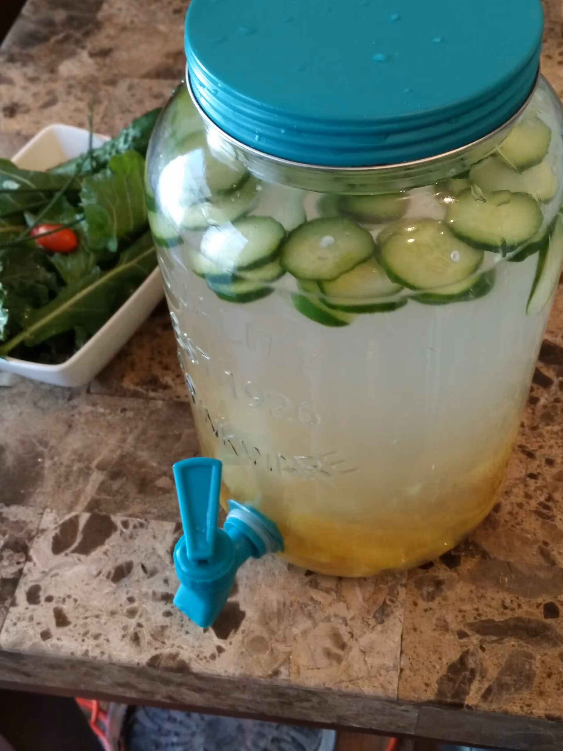 Water dispenser with cucumber slices sits on a kitchen counter beside fresh greens