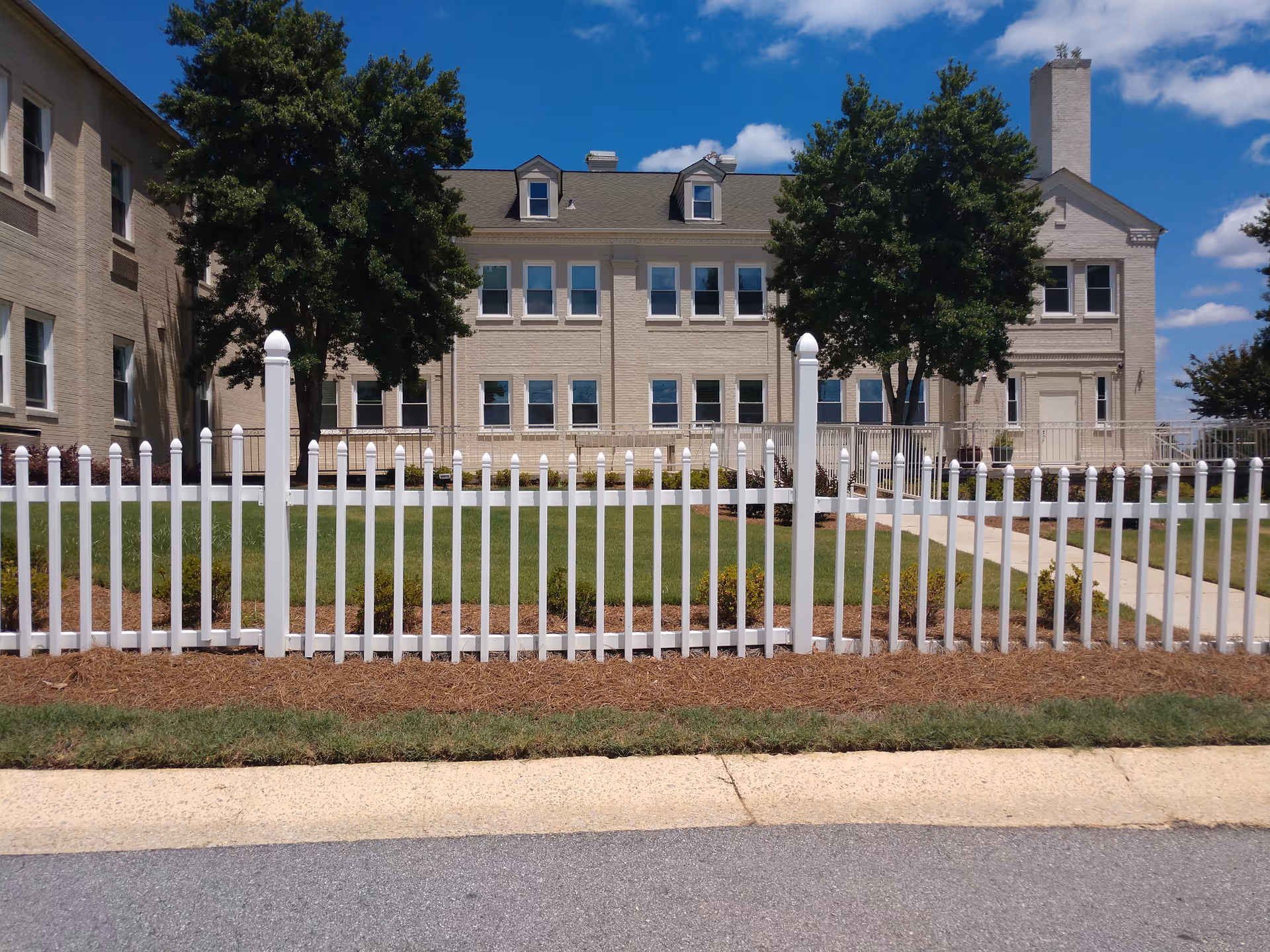 Exterior view of a beige senior living building behind a white picket fence and green lawn.