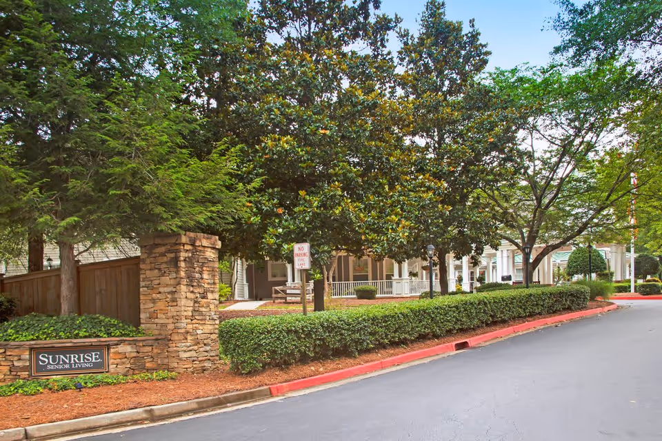 Exterior view of a senior living facility with a stone pillar and sign reading 'Sunrise Senior Living' near a driveway lined with bushes and trees. The building has a covered porch with white columns and benches.