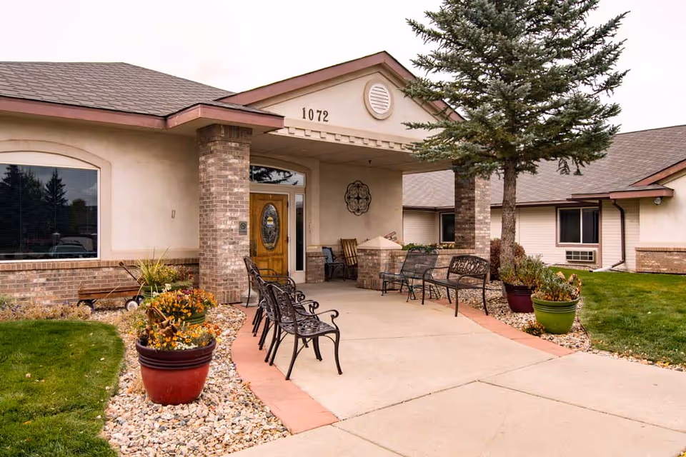 Front entrance of a single-story senior living building with benches, potted plants, and the address "1072" above the doorway.