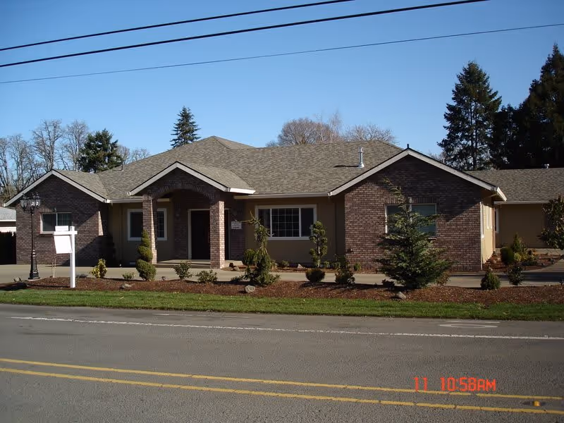 Exterior view of a single-story brick and siding building with a gabled roof, small landscaped shrubs and trees in front, and a clear blue sky above. The building appears to be a residential care home with a driveway and a street in the foreground.