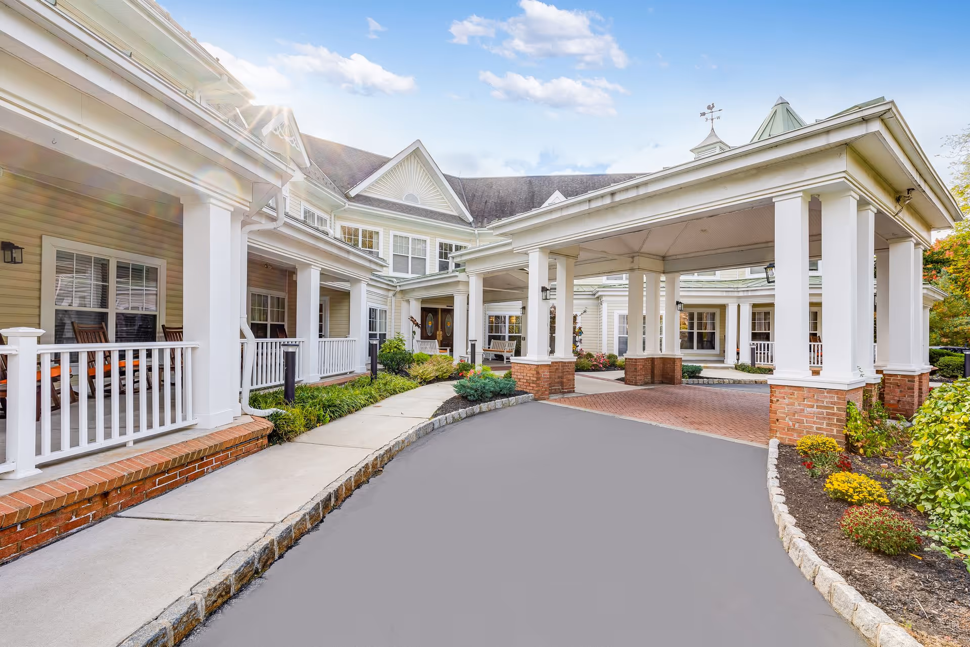 Exterior view of Sunrise of Morris Plains facility showing a covered entrance with white columns and brick bases, a driveway, landscaped garden beds with shrubs and flowers, and a porch with rocking chairs under a partly cloudy sky.