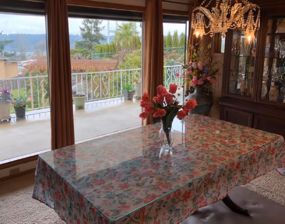 A floral tablecloth covers a dining table beneath a chandelier by large windows.