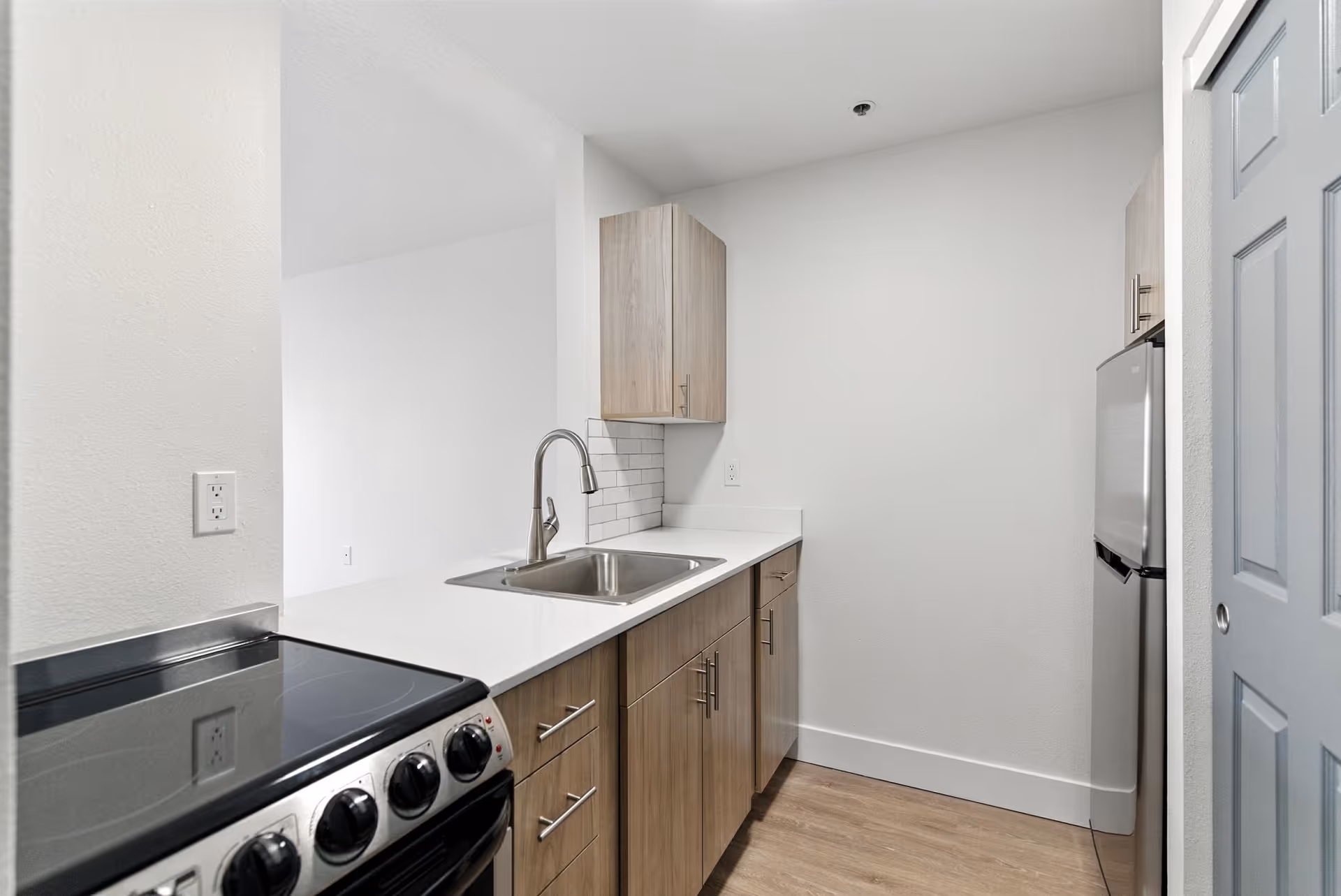 Compact modern kitchen with wood-tone cabinets, white countertops, sink, stove, and fridge.