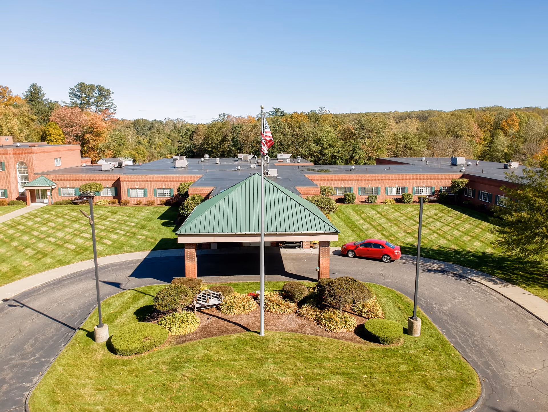 Aerial view of CareOne at Millbury facility showing a single-story brick building with a green metal roof entrance canopy. There is a circular driveway with a red car parked near the entrance. The surrounding area has neatly manicured lawns, bushes, and trees with autumn foliage in the background under a clear blue sky.