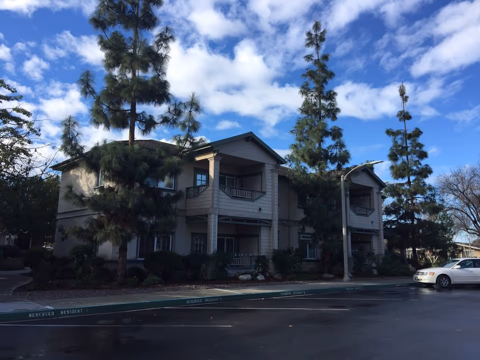 Two-story residential building with covered balconies flanked by tall pine trees under a blue sky.