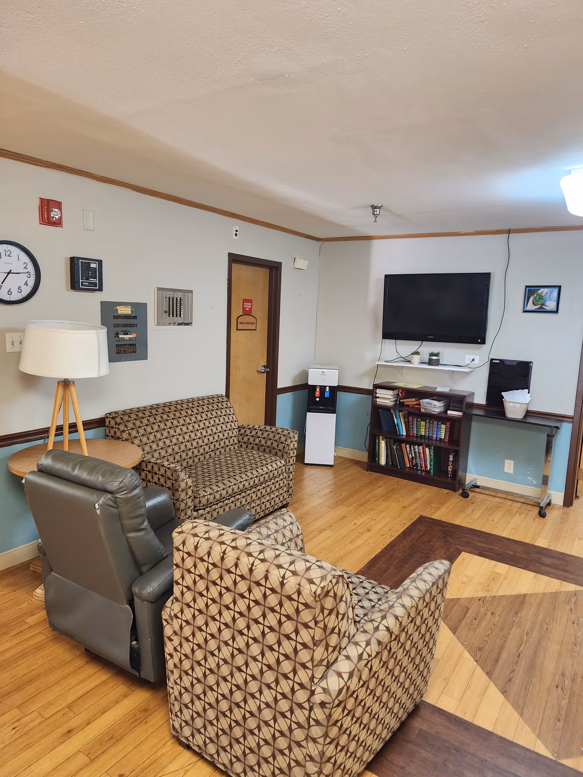 Senior living facility lounge with patterned chairs, a sofa, TV, and bookshelves.