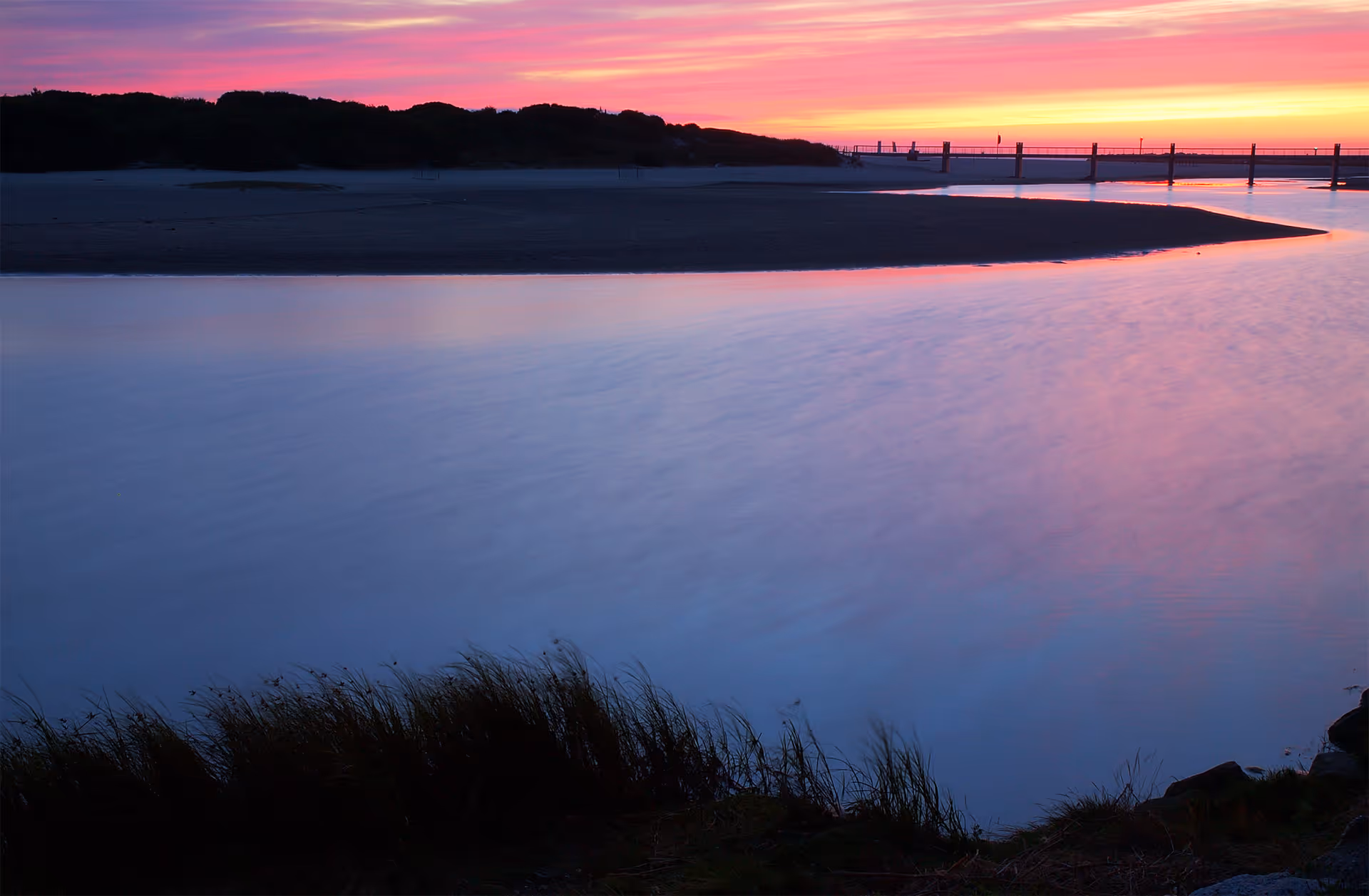Sunset over calm water with grasses in foreground and a distant pier.