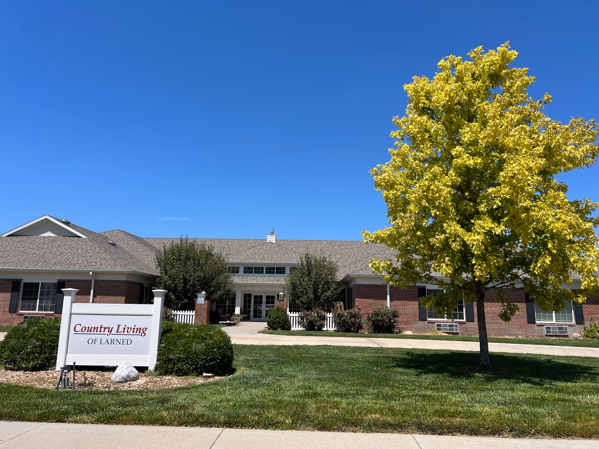 Single-story brick senior living facility with a sign, green lawn, and a yellow-leaved tree