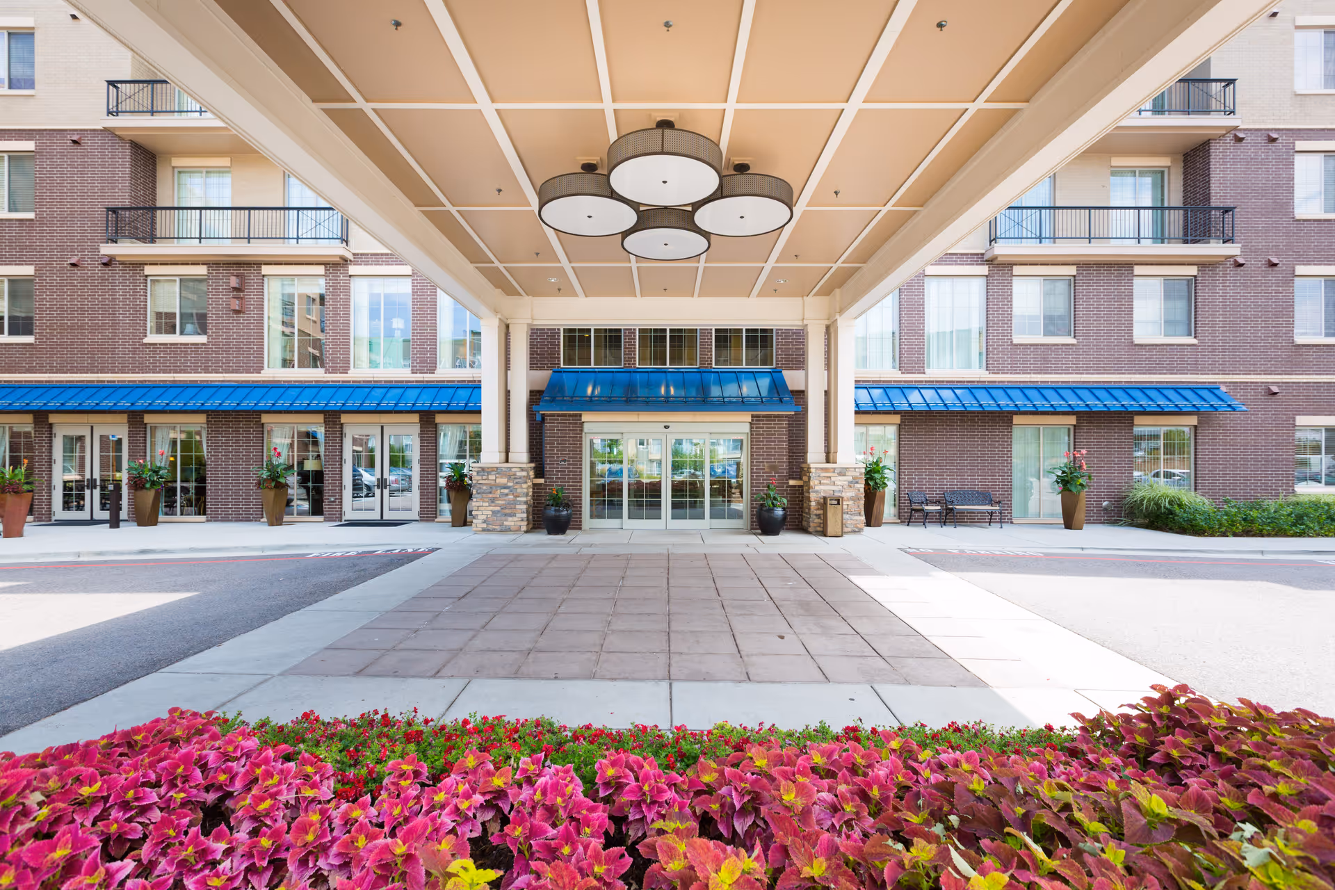 Entrance of a multi-story assisted living facility with a covered drop-off area, large glass doors, brick exterior, blue awnings, and vibrant pink and green plants in the foreground.
