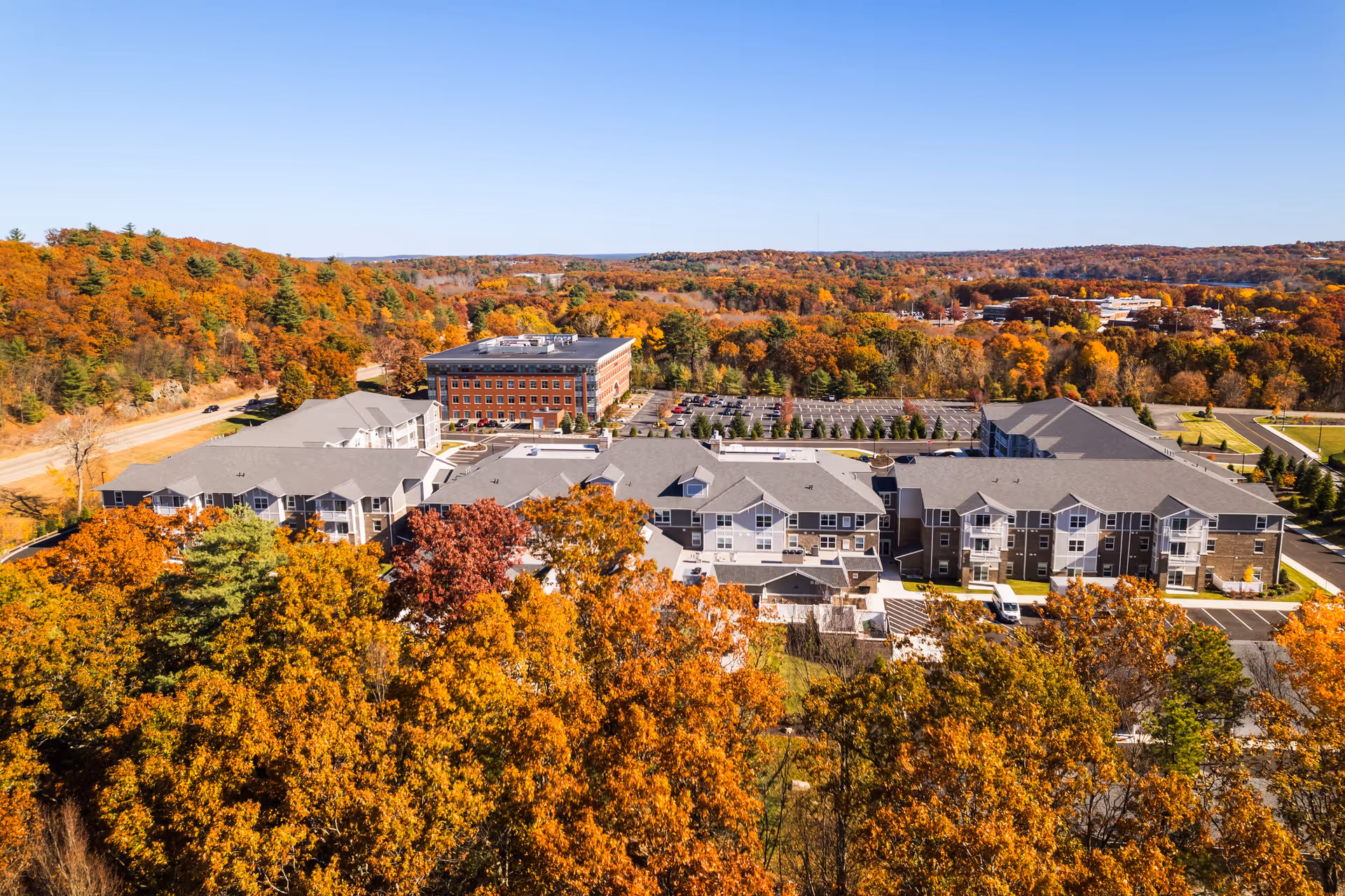 Aerial view of Pleasant Hill Retirement Community surrounded by trees with autumn foliage. The facility consists of multiple connected buildings with gray roofs and parking lots. The background shows a forested area with colorful fall trees under a clear blue sky.